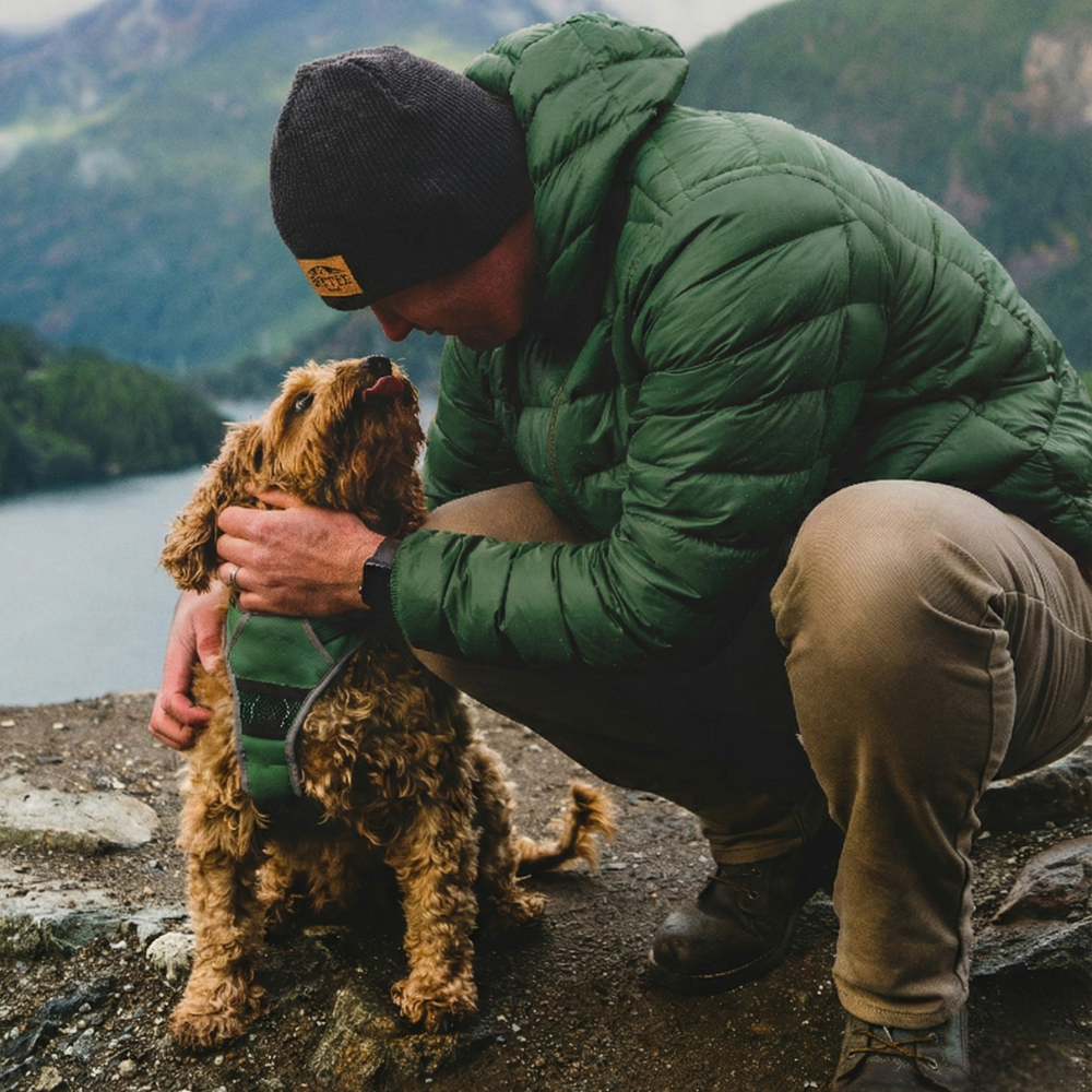 Man in green jacket and black beanie petting a dog by a lake with mountains in the background